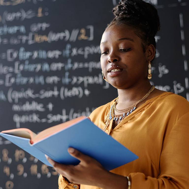 A woman stands before a blackboard, holding a book, engaged in teaching or presenting information.