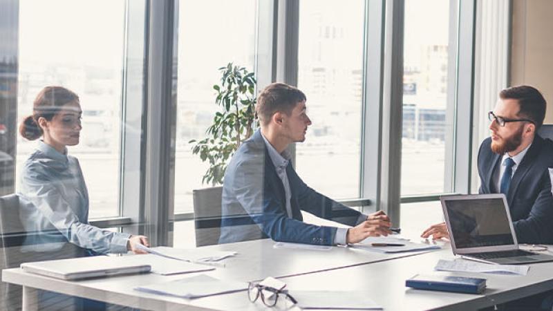 A group of business professionals engaged in discussion at a table, with a large window providing natural light behind them.