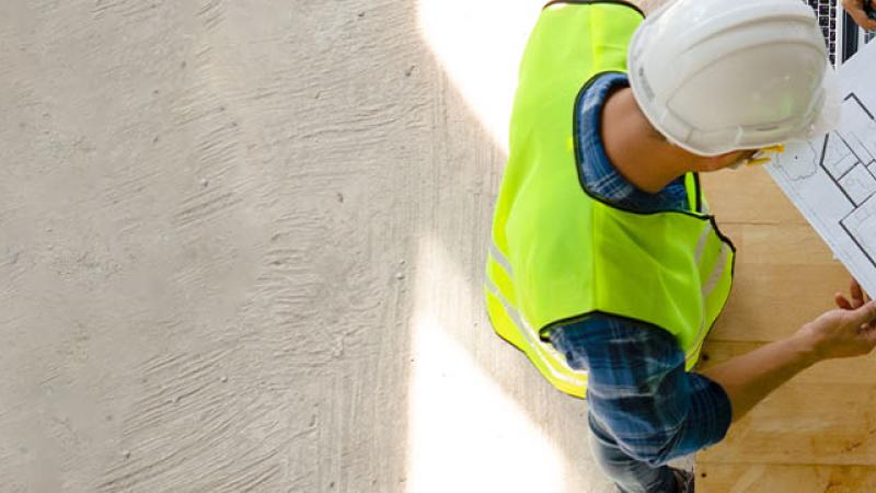 Three individuals in hard hats and safety vests examine blueprints at a construction site, focused on their task.