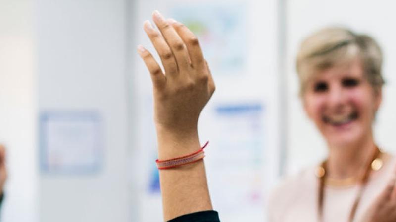 A woman stands in a classroom, raising her hands enthusiastically as she engages with her students.