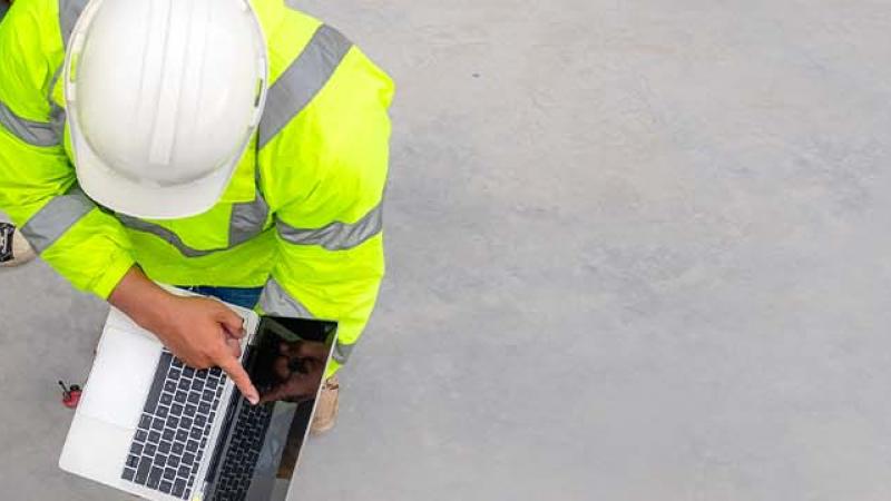 Three men in hard hats and safety vests collaborate over a laptop, focused on their work in a construction environment.