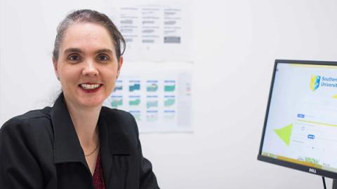 A woman seated at a desk, focused on her work in front of a computer, surrounded by office supplies and a notepad.