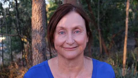 A woman with brown hair wearing a blue shirt stands amidst a backdrop of lush green trees.