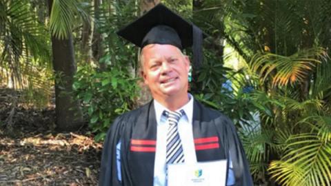 A man in a graduation gown and cap stands proudly in front of lush green trees, celebrating his academic achievement.