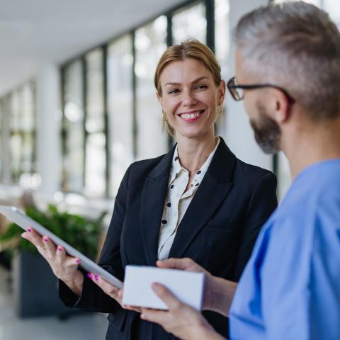 A female health professional in a business suit engages in conversation with a man, both appearing professional and focused on the discussion.