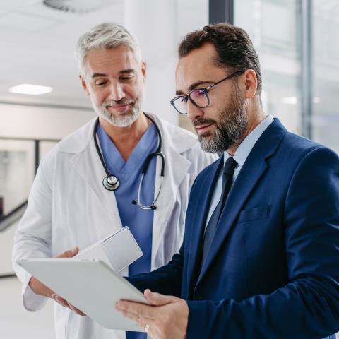 A doctor and a male MBA Health Services Management student in a suit examining a tablet together, engaged in a discussion about patient information or medical data