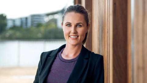 A professional woman in a suit and tie leans against a rustic wooden wall, exuding confidence and style.