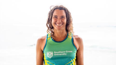 A woman wearing a green and yellow shirt stands on the beach, enjoying the sun and the ocean waves in the background.