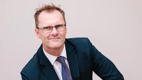 A man wearing glasses and a suit is seated on a chair, exuding a professional and composed demeanor.
