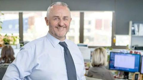 A man in a shirt and tie stands confidently in front of a computer, engaged in a professional setting.