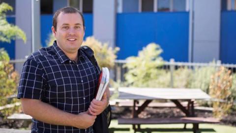 A man stands outside, smiling while holding a folder, exuding a sense of confidence and positivity.
