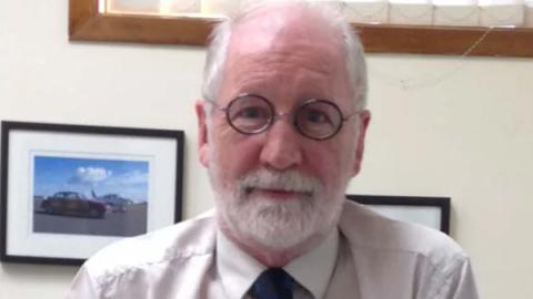 A professional woman in a suit and tie leans against a rustic wooden wall, exuding confidence and style.