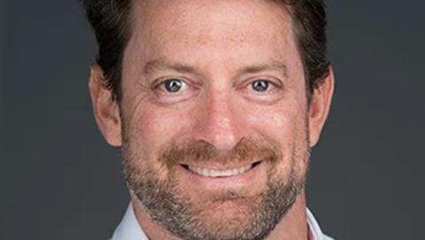 A bearded man wearing a white shirt, standing confidently with a neutral expression in a well-lit environment.