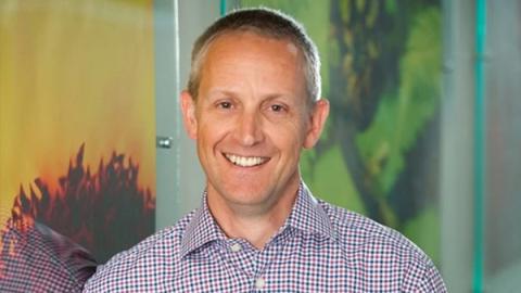 A man wearing a checkered shirt smiles warmly at the camera, exuding a friendly and approachable demeanor.