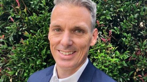 A man in a suit and tie smiles confidently while standing in front of lush green bushes.