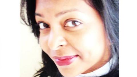 A woman with dark hair wearing a white shirt, standing confidently with a neutral expression against a plain background.