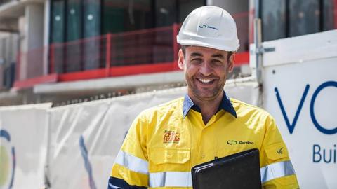 A man wearing a yellow hard hat is holding a laptop, focused on his work in a construction or engineering setting.
