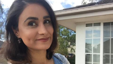 A woman with dark hair wearing a blue top, smiling and standing against a neutral background.