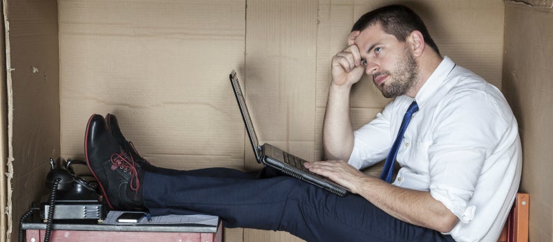 A man sat on a desk looking stressed with a laptop. 